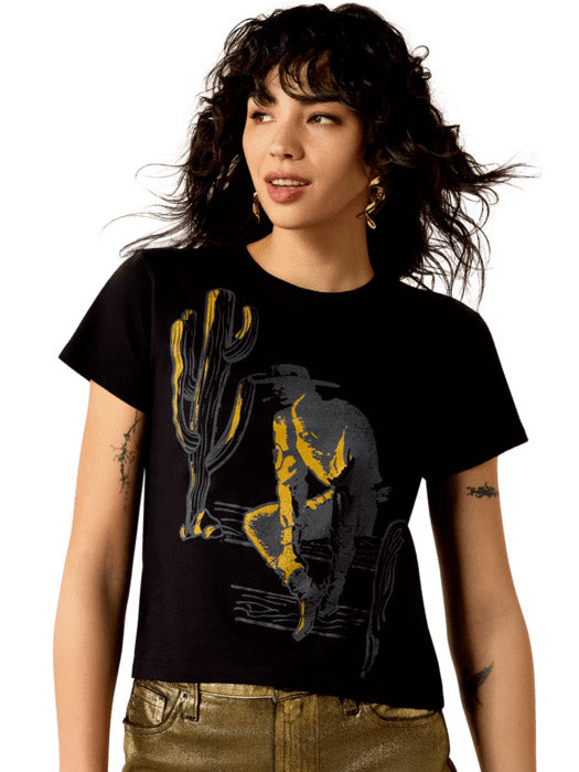 A woman modeling a black short-sleeve graphic t-shirt with a yellow and gray desert cowboy design, paired with metallic gold pants, against a plain white background. If you need any assistance with this item or the purchase of this item please call us at five six one seven four eight eight eight zero one Monday through Saturday 10:00a.m EST to 8:00 p.m EST