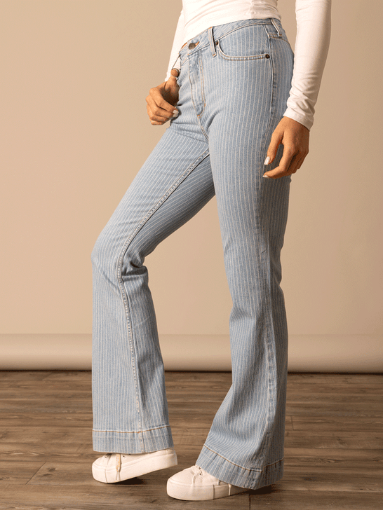 Person wearing light blue flared jeans on a wooden floor with a beige wall background side view. If you need any assistance with this item or the purchase of this item please call us at five six one seven four eight eight eight zero one Monday through Saturday 10:00a.m EST to 8:00 p.m EST